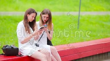两个女孩在户外使用智能手机。 两个女人坐在公园里，用智能手机发送信息